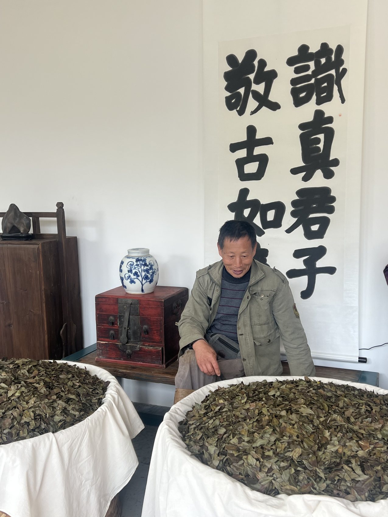 Ernan’s father seated among trays of withering leaf beneath calligraphy scrolls